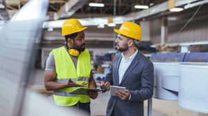 Zwei Männer mit gelben Schutzhelmen stehen in einer Industriehalle.