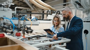 Zwei Fachkräfte mit Schutzbrillen prüfen eine industrielle Produktionsanlage und dokumentieren die Ergebnisse digital auf einem Tablet.