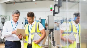Zwei Männer in einer Produktionshalle besprechen Informationen auf einem Tablet. Im Hintergrund sind Maschinen und Arbeitsgeräte sichtbar.