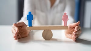 Zwei Figuren – eine blau, eine rosa – stehen auf einer Waage und symbolisieren Gleichgewicht und Gleichbehandlung, während eine Person die Balance hält.