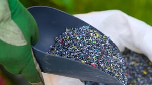 Nahaufnahme einer Schaufel mit buntem Kunststoffgranulat in einer Hand mit grünem Arbeitshandschuh.