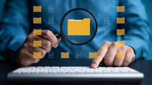 Person hält eine Lupe über einem digitalen Ordnersymbol, umgeben von weiteren Datei- und Ordnericons.