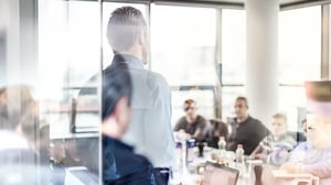 Blick durch eine Glaswand in einen modernen Konferenzraum, in dem ein Mann vor einer Gruppe steht und spricht.
