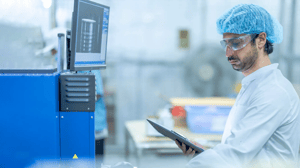 Mitarbeitender in Schutzkleidung und Schutzbrille arbeitet in einer Produktionsumgebung mit Tablet und überwacht Prozesse.