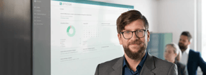 Mann steht vor großem Bildschirm mit Dashboard der Compliance Management Software Eticor, zwei Personen im Hintergrund