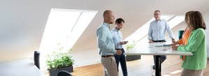 Fünf Personen stehen an einem hohen Besprechungstisch im modernen, lichtdurchfluteten Büro und tauschen sich aus.