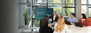 Frau präsentiert Workshop-Folie, vier Personen sitzen mit Laptops am Tisch und hören zu.