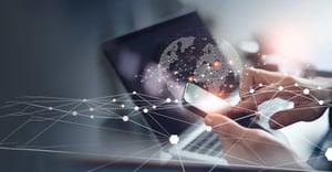 Hände bedienen ein Smartphone vor einem Laptop, darüber eine digitale Weltkugel mit vernetztem Datenfluss – Symbolbild für globale digitale Kommunikation.