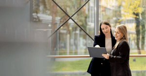 Zwei Frauen betrachten gemeinsam ein Tablet, im Hintergrund ein verglaster Büroflur mit Blick ins Grüne.