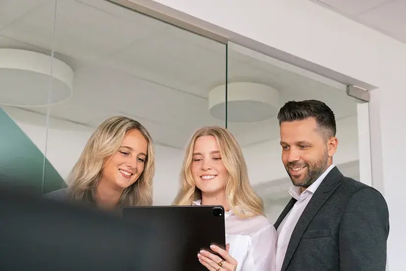 Drei Personen schauen in einem modernen Büro mit Glaswand lächelnd auf ein Tablet. 