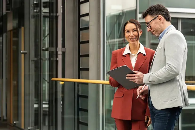 Zwei Personen im Gespräch in einem modernen Bürogebäude mit Glasfassade, in der Hand hält der Mann ein Tablet.