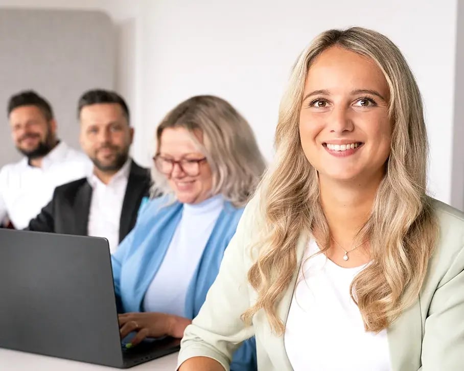 Junge Frau lächelt in die Kamera, im Hintergrund Teammitglieder in einem Meeting.