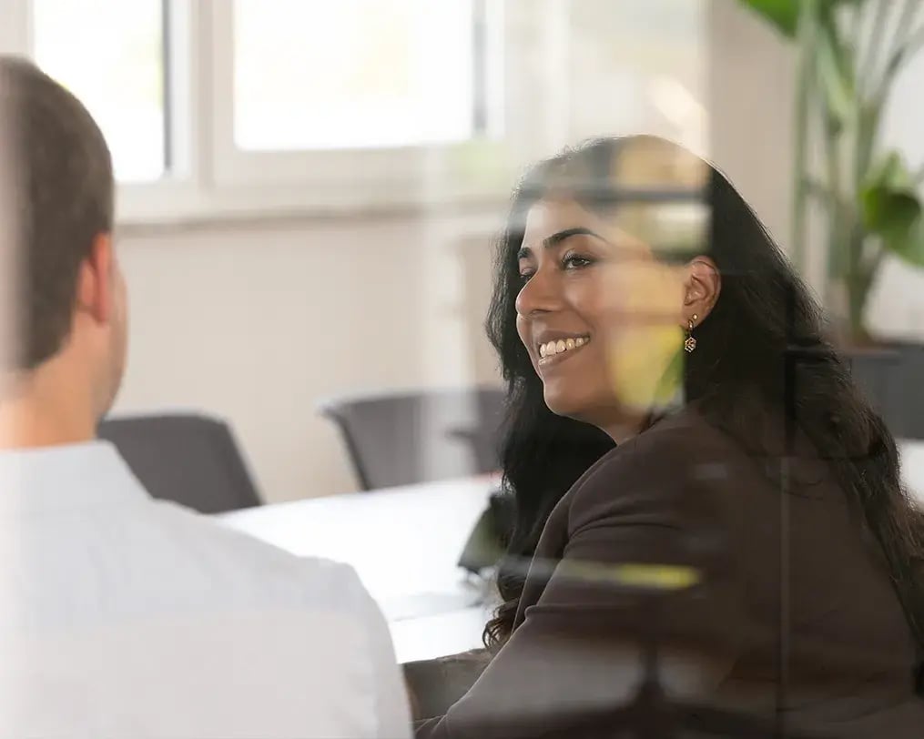 Zwei Personen im Gespräch, eine Frau blickt lächelnd durch eine Glasscheibe auf ihren Gesprächspartner. Zwei Personen im Gespräch, eine Frau blickt lächelnd durch eine Glasscheibe auf ihren Gesprächspartner.
