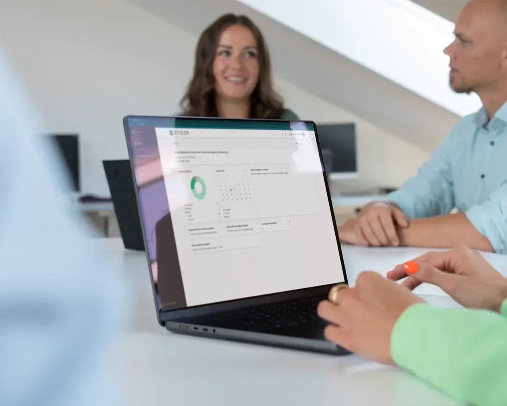 Laptop mit geöffneter Eticor Compliance Management Software auf einem Konferenztisch, im Hintergrund mehrere Personen.