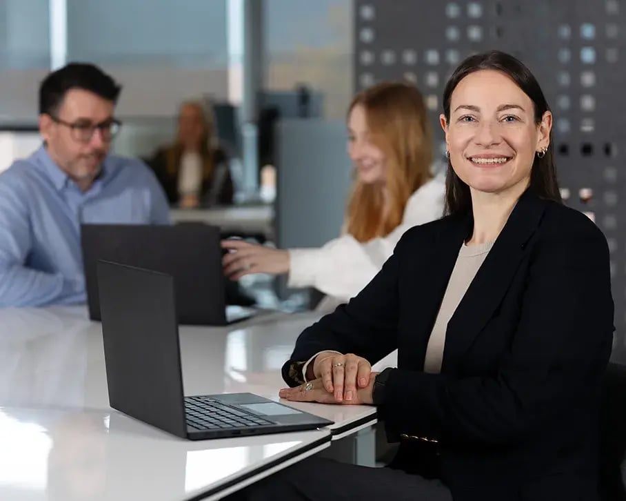 Frau am Besprechungstisch mit Laptop, im Hintergrund zwei weitere Teammitglieder bei der Arbeit. Frau am Besprechungstisch mit Laptop, im Hintergrund zwei weitere Teammitglieder bei der Arbeit.