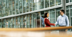 Two people shaking hands and smiling in front of a glass office building.