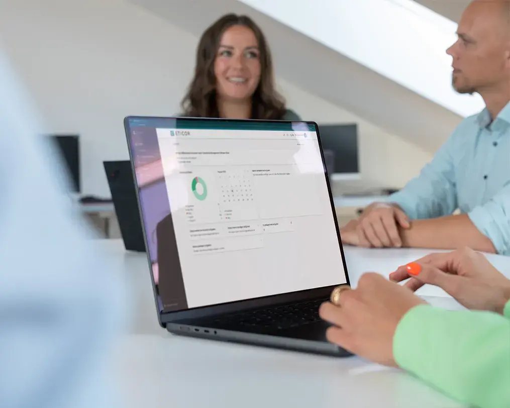 Laptop mit Eticor Compliance Management Software auf einem Konferenztisch, im Hintergrund sprechen mehrere Personen.