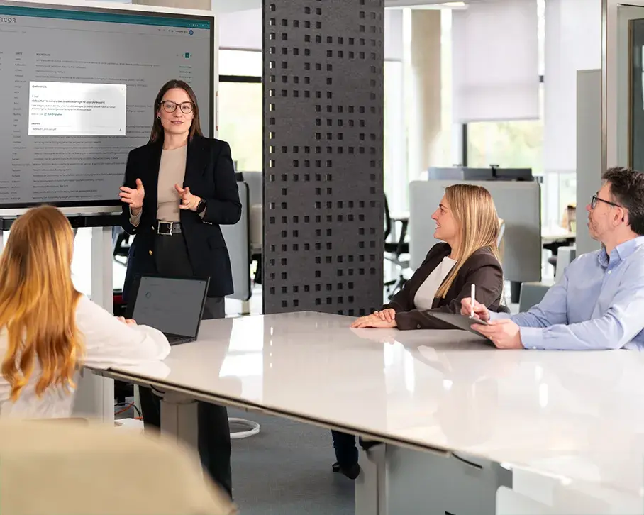 Frau präsentiert Eticor-Software auf großem Bildschirm in Meetingraum, drei Personen folgen der Präsentation.